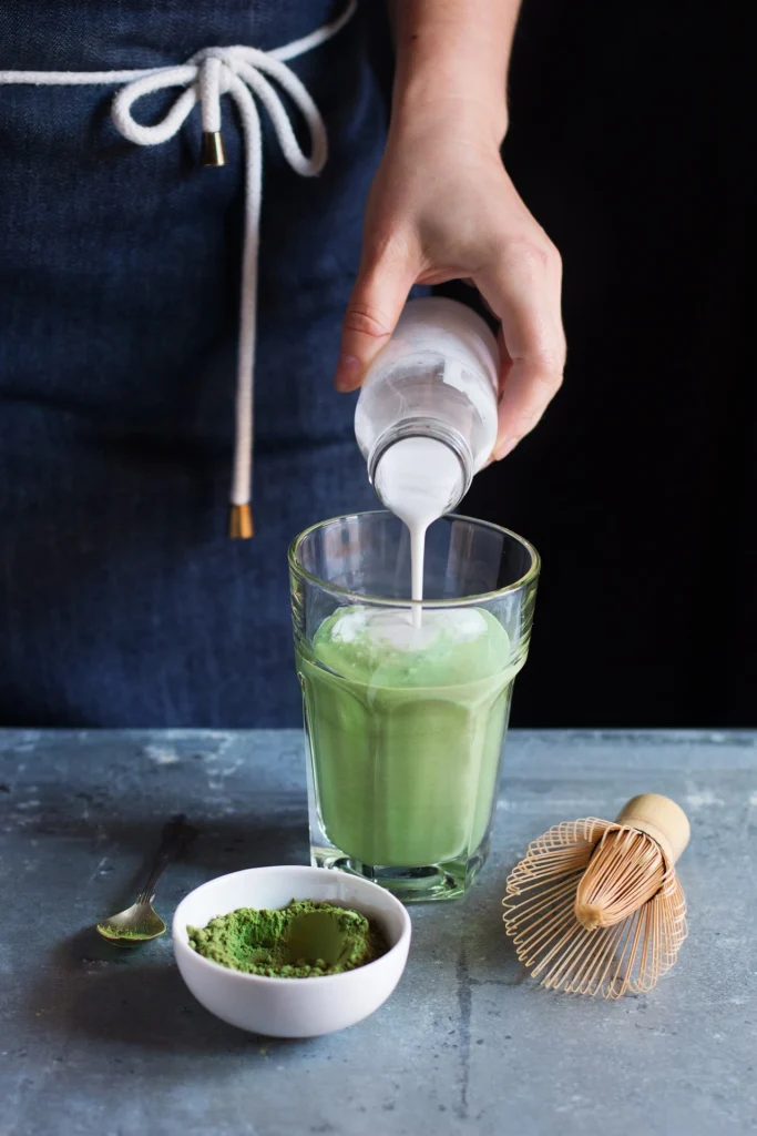 Person gießt Milch in ein Glas mit Matcha-Latte, daneben Matcha-Pulver und ein Bambusbesen auf einer dunklen Oberfläche